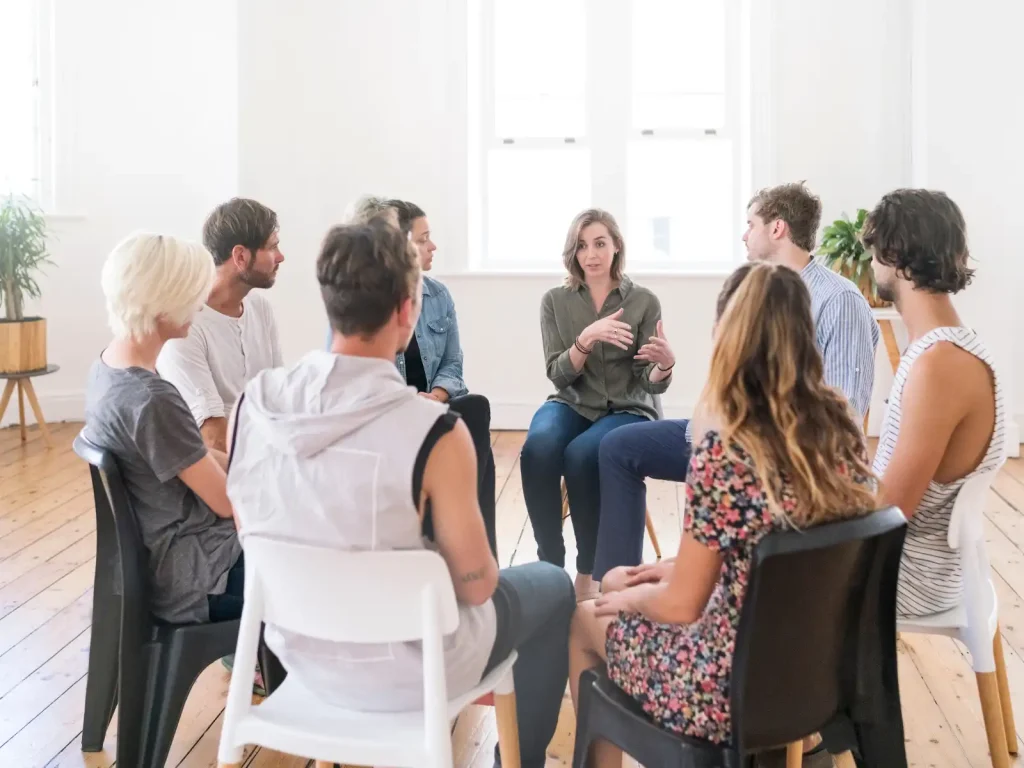 A supportive ADHD couples support group gathered in a bright, welcoming space. A woman leads the discussion while others listen attentively, fostering connection and understanding.