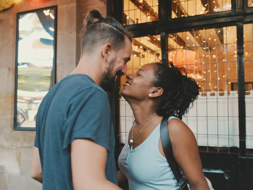 A happy couple shares a loving moment outside a warmly lit café, their faces close as they smile at each other. String lights reflect in the window behind them, creating a cozy and intimate atmosphere.