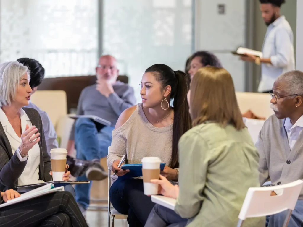 A diverse group of individuals engaged in a supportive group therapy session, attentively listening and sharing their experiences in a welcoming environment.