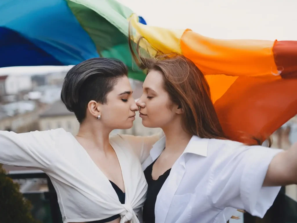 A loving LGBTQ+ couple stands close, embracing under a flowing rainbow flag. A powerful representation of acceptance and support in therapy with Grazel Garcia and Associates.