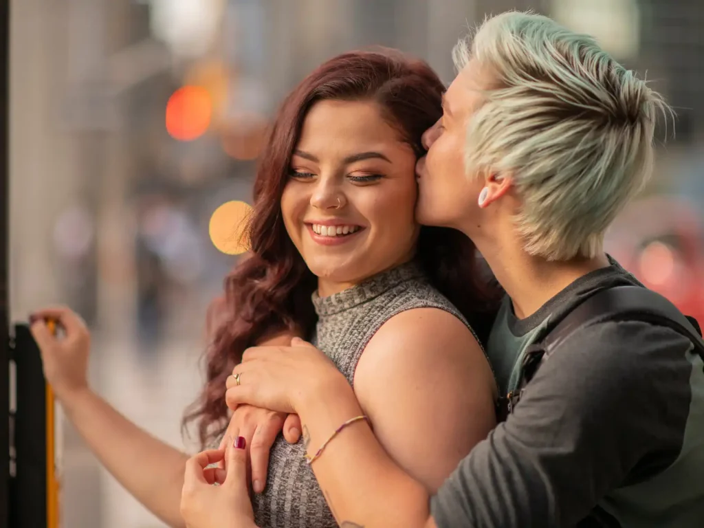 A joyful couple in an intimate embrace, with one partner kissing the other's cheek in a warm city setting, symbolizing love, connection, and support for LGBTQ+ relationships.