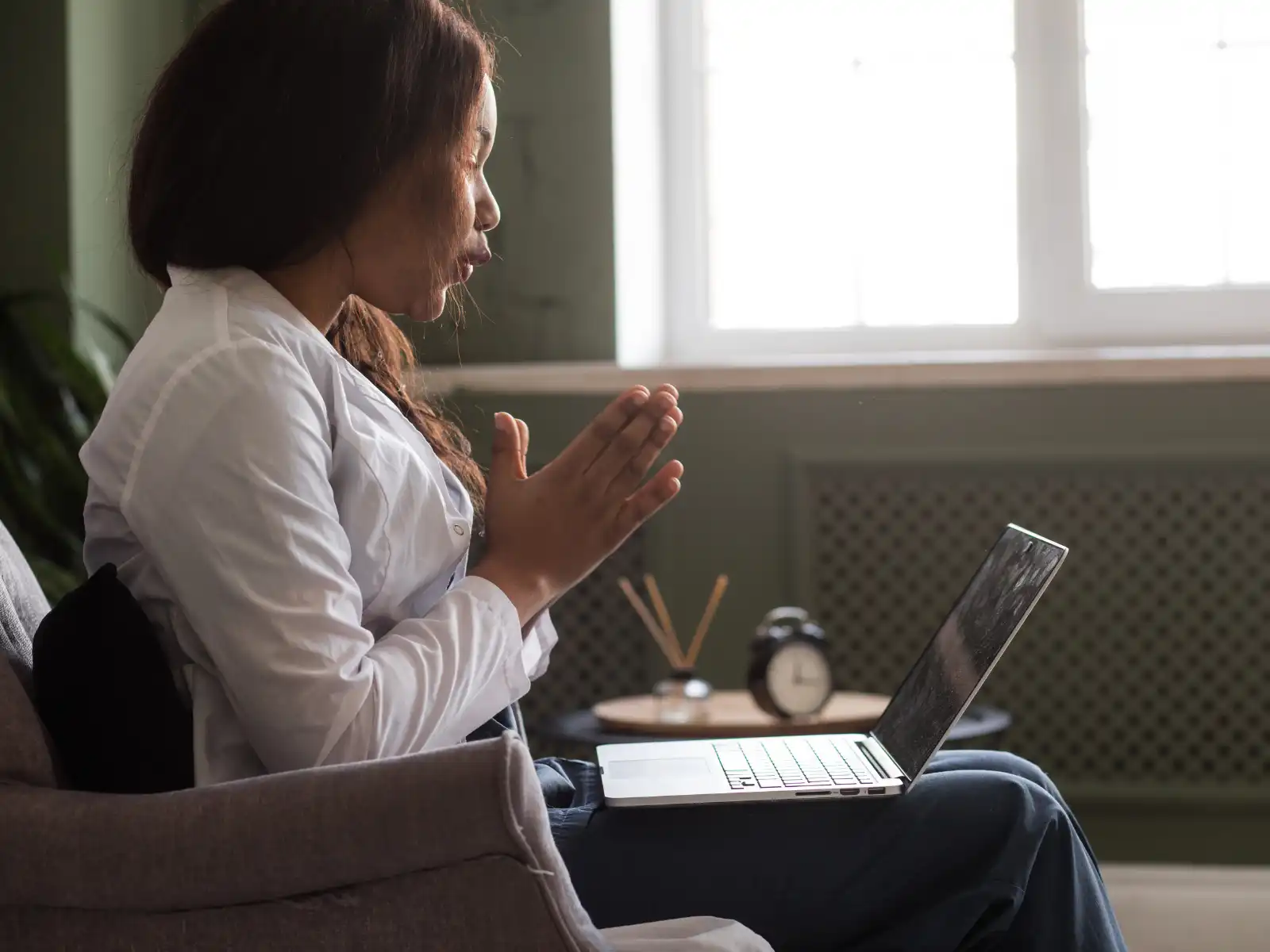 A therapist in a white coat engages in an online therapy session via laptop. She sits in a cozy, well-lit room, speaking attentively with her hands clasped, conveying empathy and professionalism.
