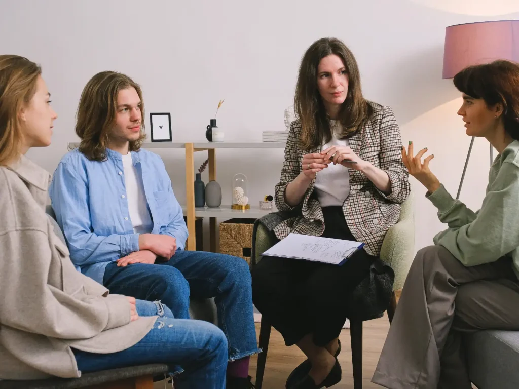 A group therapy session in progress, featuring a professional therapist leading a discussion with three individuals in a calming and supportive environment.