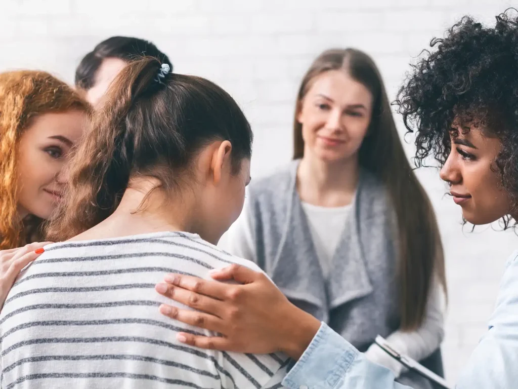 A supportive therapy group session where a young woman receives comforting gestures from fellow members