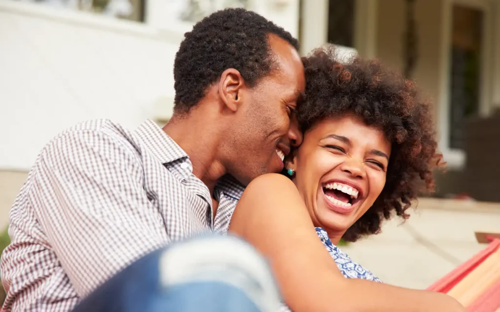 Co-regulating-couple-therapy A black man holding a black woman on the ground in a romantic embrace. Both people are smiling and laughing demonstrating good co-regulation after successful couple therapy