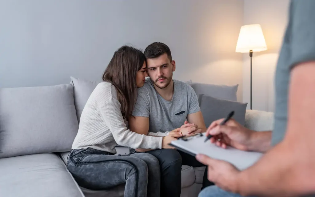 Couples-therapy-session A white man and woman sitting on a therapist couch during a couples therapy session. The woman is cuddling up to the man, holding his hand and resting her chin on her shoulder.