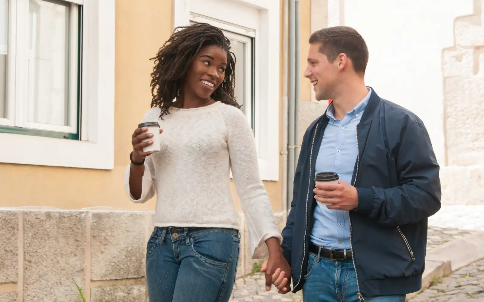 An interracial couple walking down a cobbled street, holding cups of coffee and each others' hands, smiling at each other