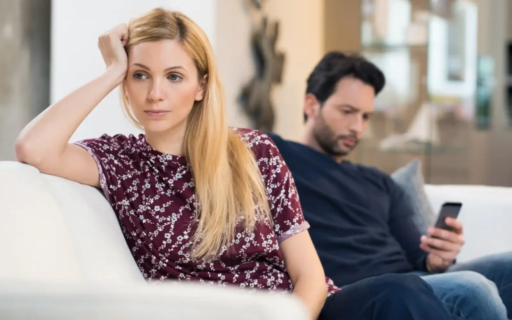 Dismissing-concerns-around-microaggressions An interracial couple sitting on a sofa. The woman is turned away from the man, who is on his phone, after he dismissed her concern around experiencing racial microaggressions