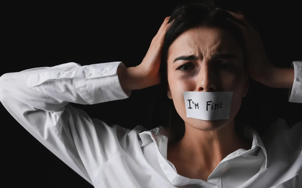 Domestic-violence-training-therapists A stressed woman with her hands on her head pulling her hair out with a sticker over her mouth that reads "I'm fine", symbolizing the impact of domestic violence on the Victim
