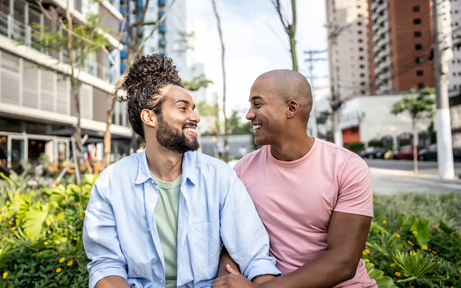 A gay interracial couple smiling and talking to each other on a bench in a city