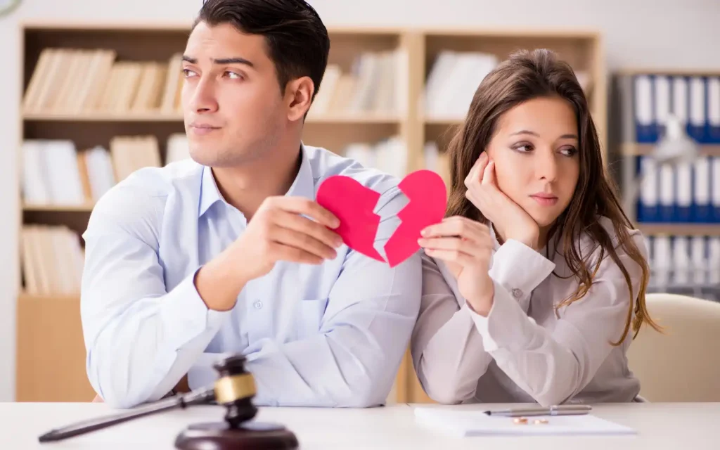 Interracial-couples-therapy-needed An interracial couple sitting at a desk facing away from each other holding a broken paper heart, representing the importance of undergoing interracial couples therapy early