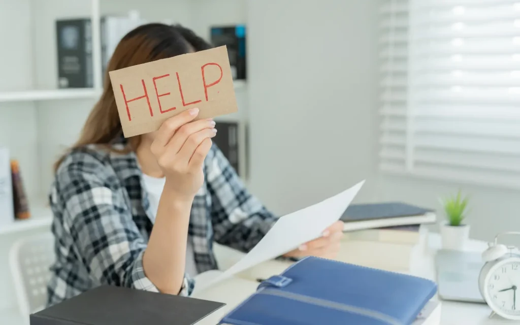 Relationship-difficulties A woman sitting at a desk holding up a piece of paper with the word help written on it representing relationship difficulties experience by neurodivergent to couples