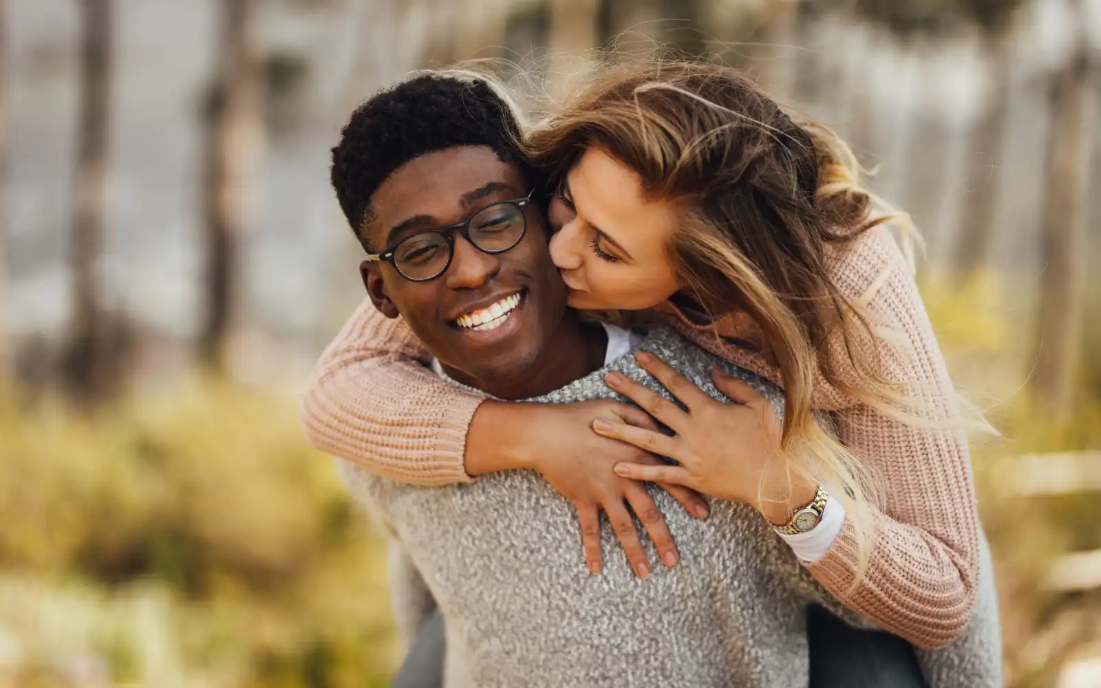 A smiling black man carrying a white woman on his back through a secluded wood. The woman is kissing the man on the cheek and the man is smiling, representing how couples want their relationships to be after couples therapy