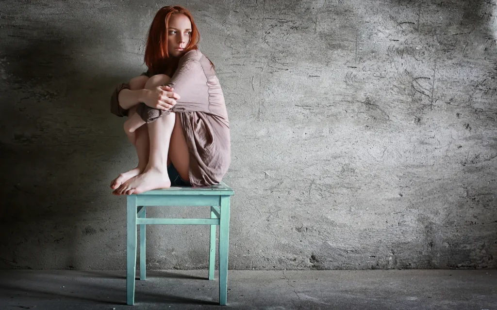 Trauma-in-relationships A woman in front of a gray wall sitting on a dirty wooden chair with her knees pulled up to her chest and her arms wrapped around her legs, symbolizing the impact of trauma in relationships