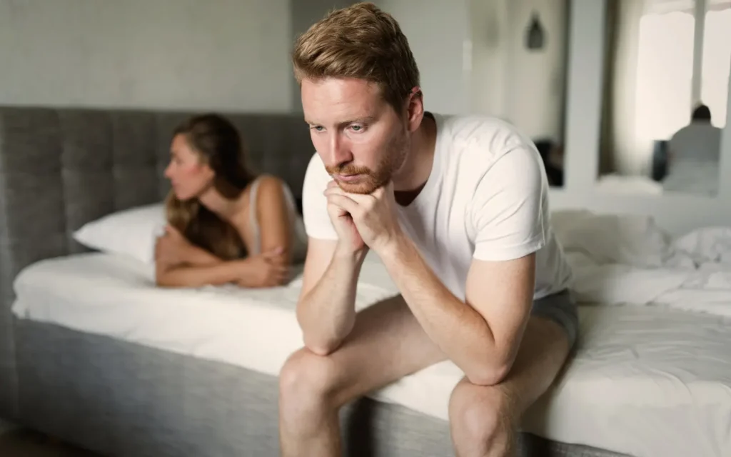 Couple-hurting-after-cheating A man sitting on the end of a bed and a woman lying on the bed, both looking distressed, representing the hurt caused by cheating