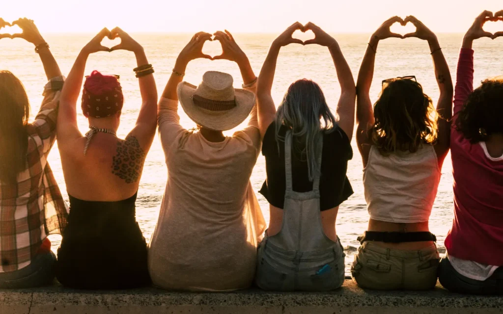 An interracial mix of women sitting on a bench looking out to the Pacific with their hands above their heads making the shape of a heart
