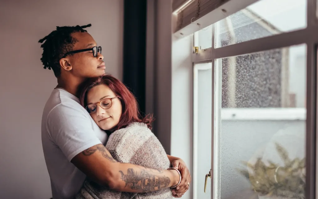 A black man and white woman embracing in an intimate hug beside a rain-speckled window, representing the safe space that interracial couples therapy can provide