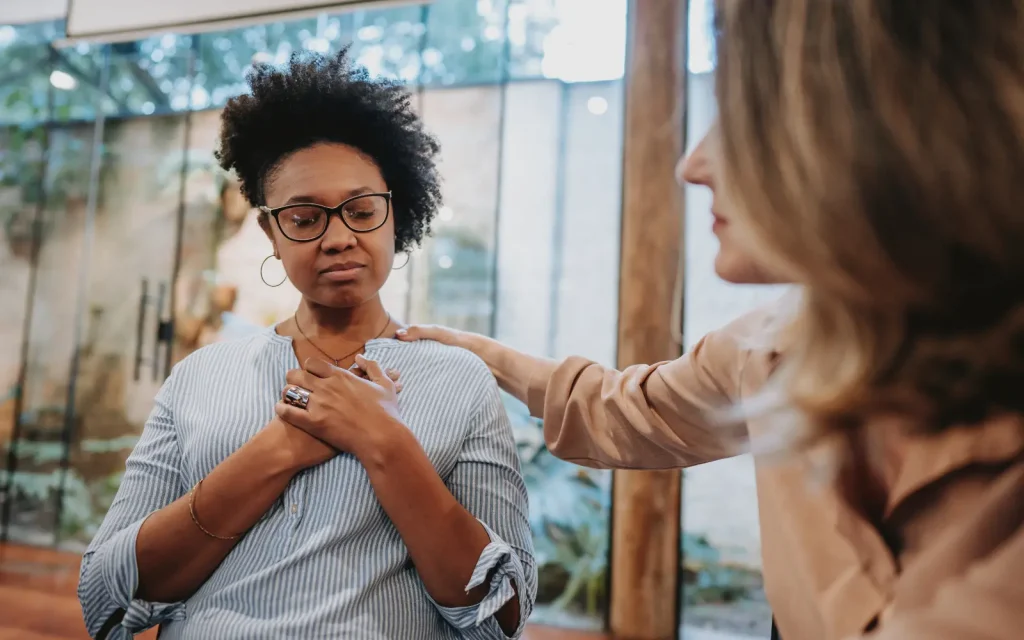 Emotional-safety-in-therapy A therapist places a comforting hand her client's shoulder while the client places her hands over her heart in a posture of gratitude, representing emotional safety in individual therapy sessions