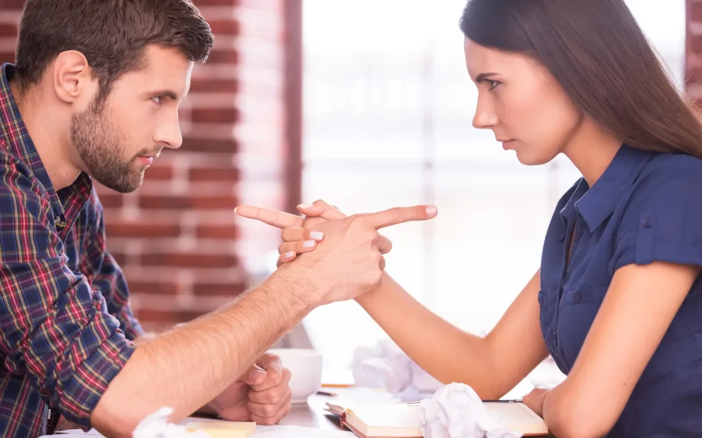 Defensive-couples-in-couples-therapy-LA A heterosexual couple facing each other across a table and pointing fingers at each other, representing couples getting defensive in couples therapy