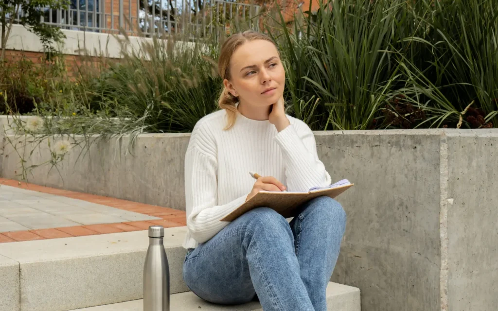 Self-reflection-through-therapy A woman sitting on steps outside looking pensive and holding a journal, representing the self-reflection that's possible through therapy