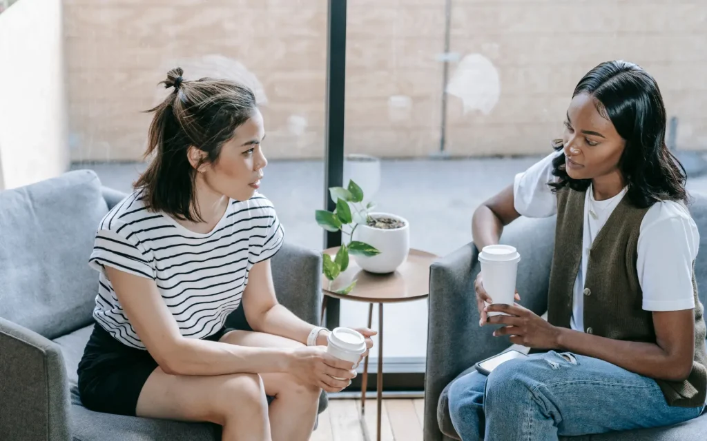 Two women sitting opposite each other holding coffee, discussing difficult topics after a marriage counseling session