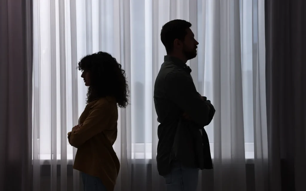 A couple in a darkened room facing away from each other with arms crossed, representing signs you may need marriage counseling