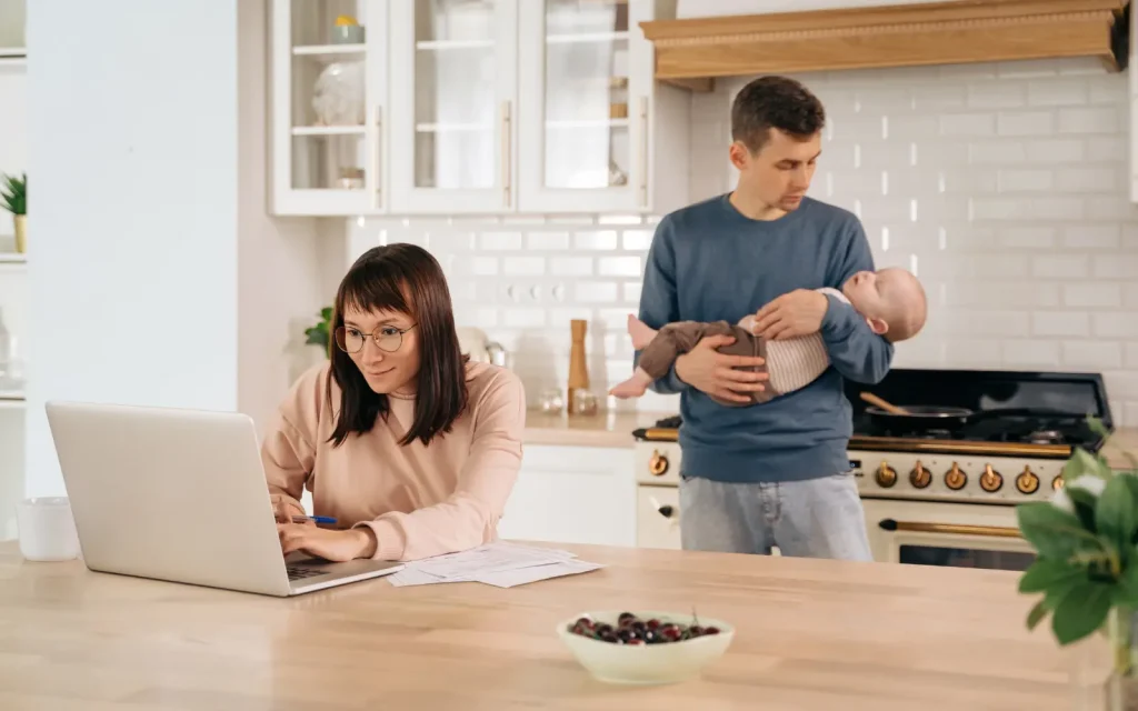 Gender-roles-in-couples-therapy A women working on a laptop while her husband bounces a baby, representing flipped gender roles that need to be discussed in pre-marital counseling in LA