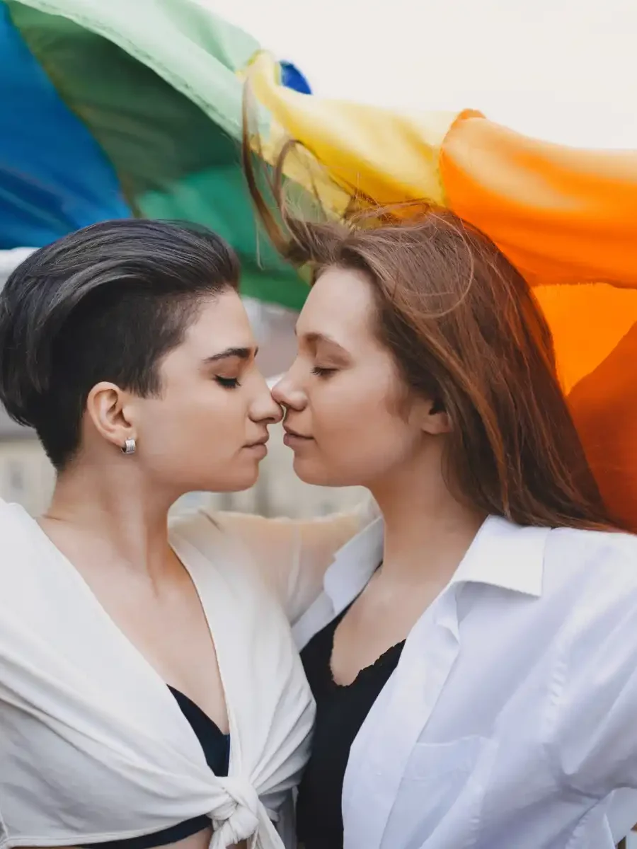 A loving LGBTQ+ couple stands close, embracing under a flowing rainbow flag. A powerful representation of acceptance and support in therapy with Grazel Garcia and Associates.