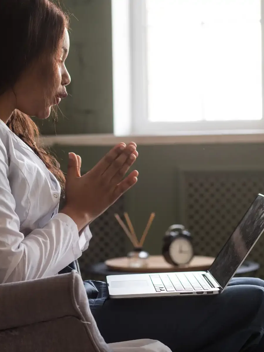 A therapist in a white coat engages in an online therapy session via laptop. She sits in a cozy, well-lit room, speaking attentively with her hands clasped, conveying empathy and professionalism.